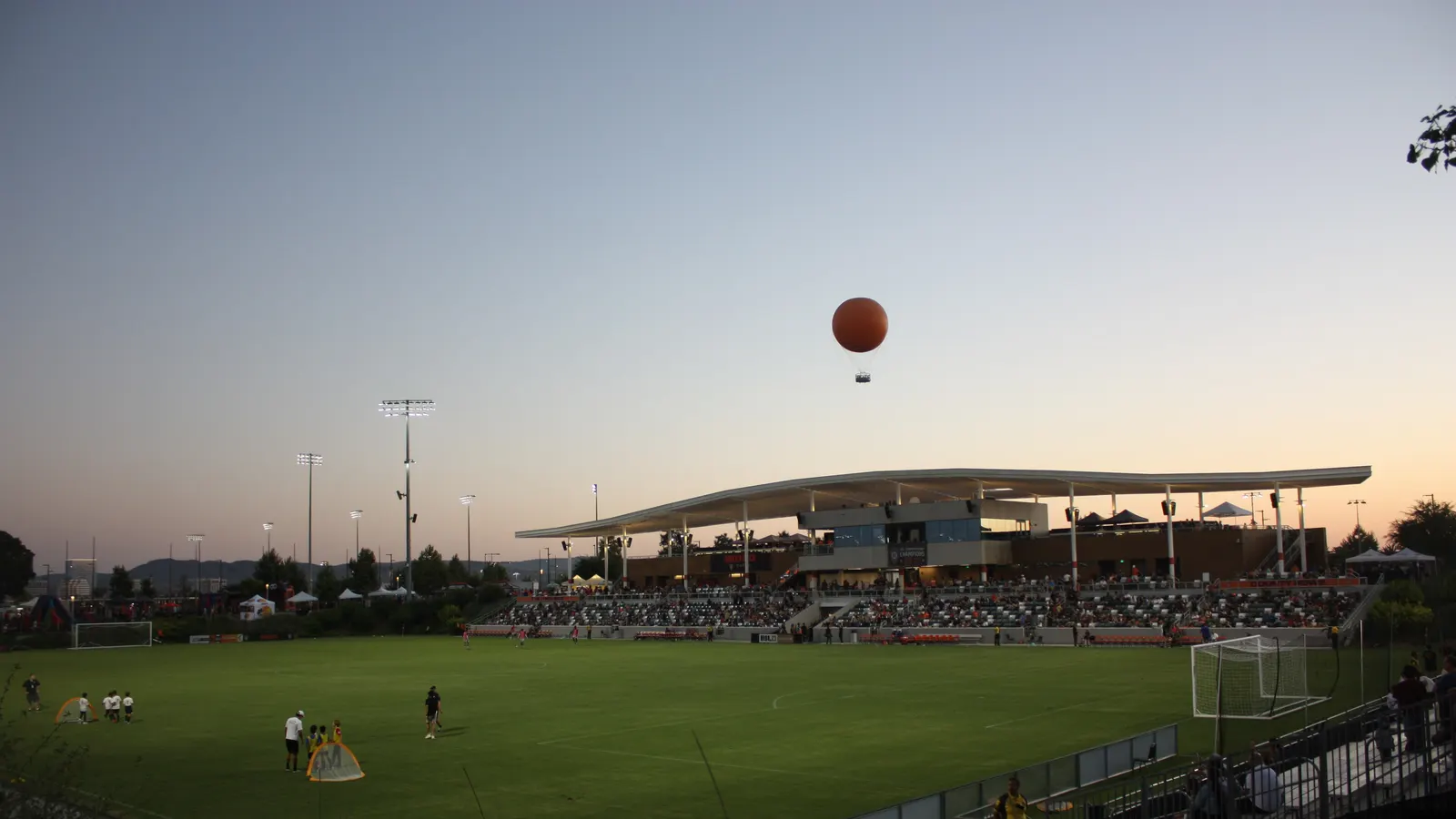 Petit stade de soccer au crépuscule avec une tribune couverte, des projecteurs et une montgolfière au-dessus du terrain