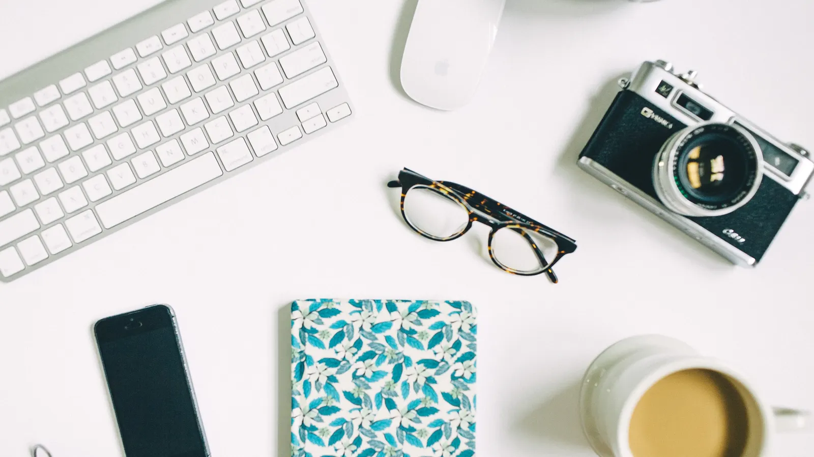 Bureau vu du dessus avec un clavier, un appareil photo, des lunettes, un téléphone, un café et un carnet fleuri